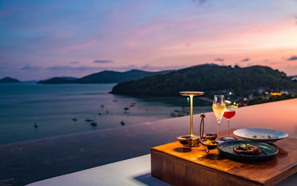 A table set with cocktails and small plates above a Thai bay at dusk