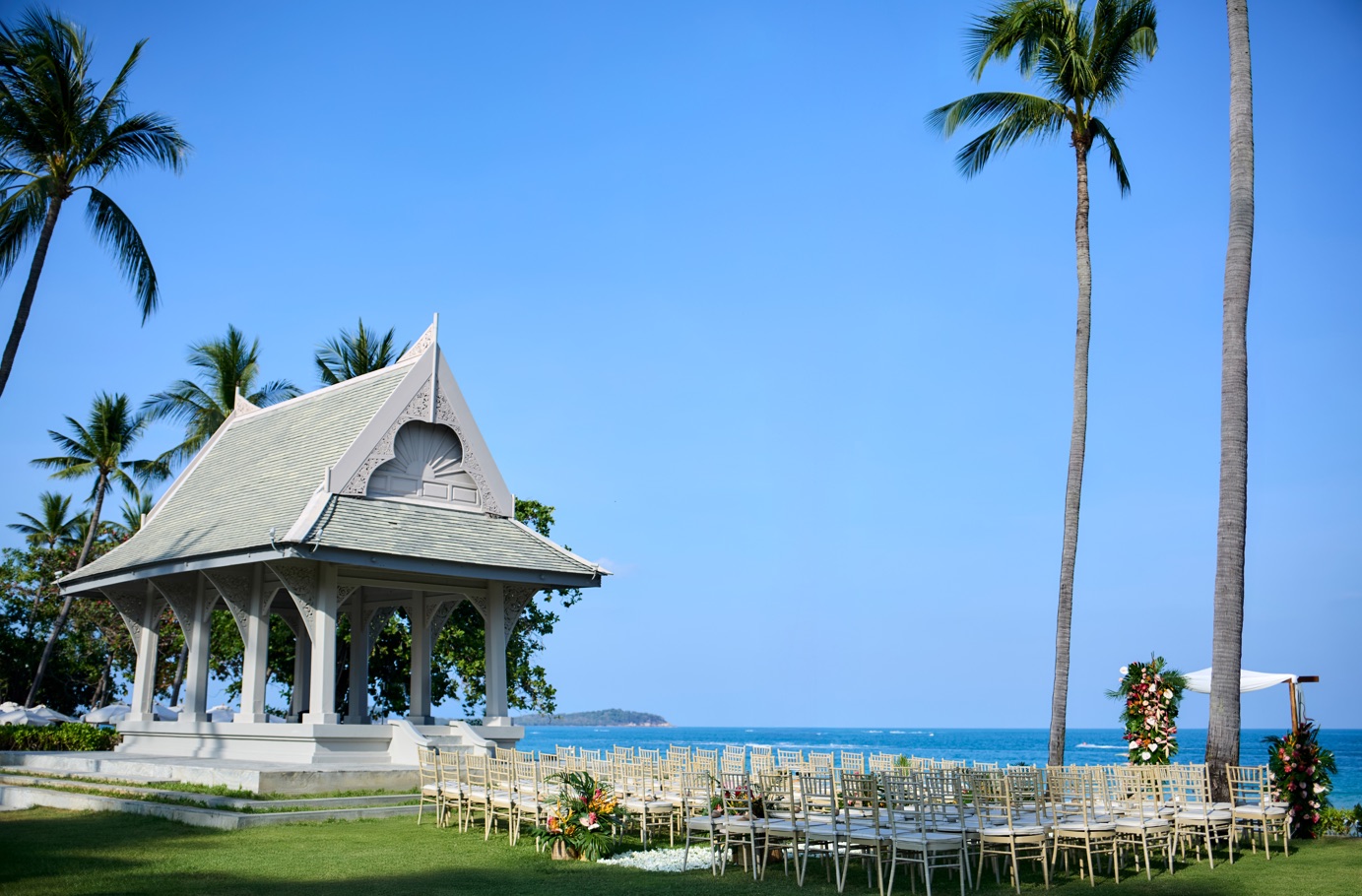 The white Thai sala pavilion on the lawn at Centara Reserve Samui, set with chiavari chairs and a floral arch for a wedding ceremony above the sea