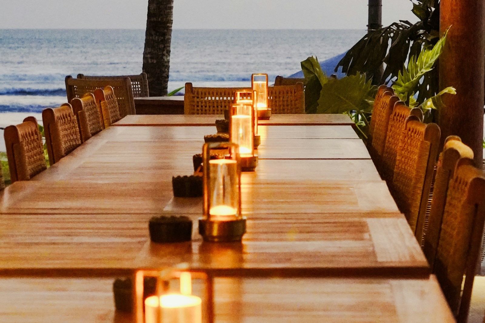 A long candle-lit table with wicker chairs set beside the sea at dusk, with a palm trunk and tropical plants at its far end
