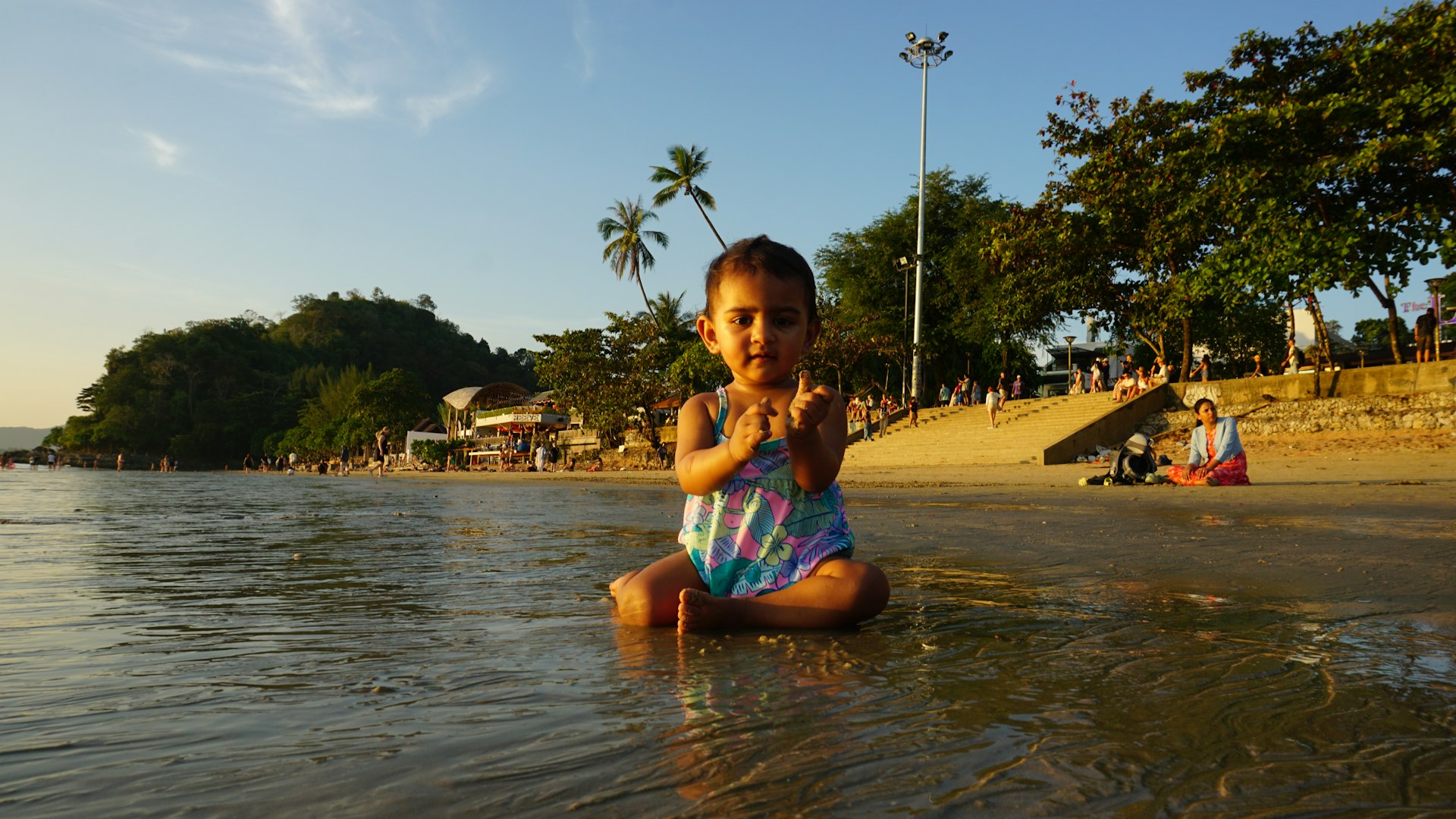 A toddler in a striped swimsuit sitting in calm, ankle-deep water at a tropical beach in the late afternoon sun, with palms and a green headland behind